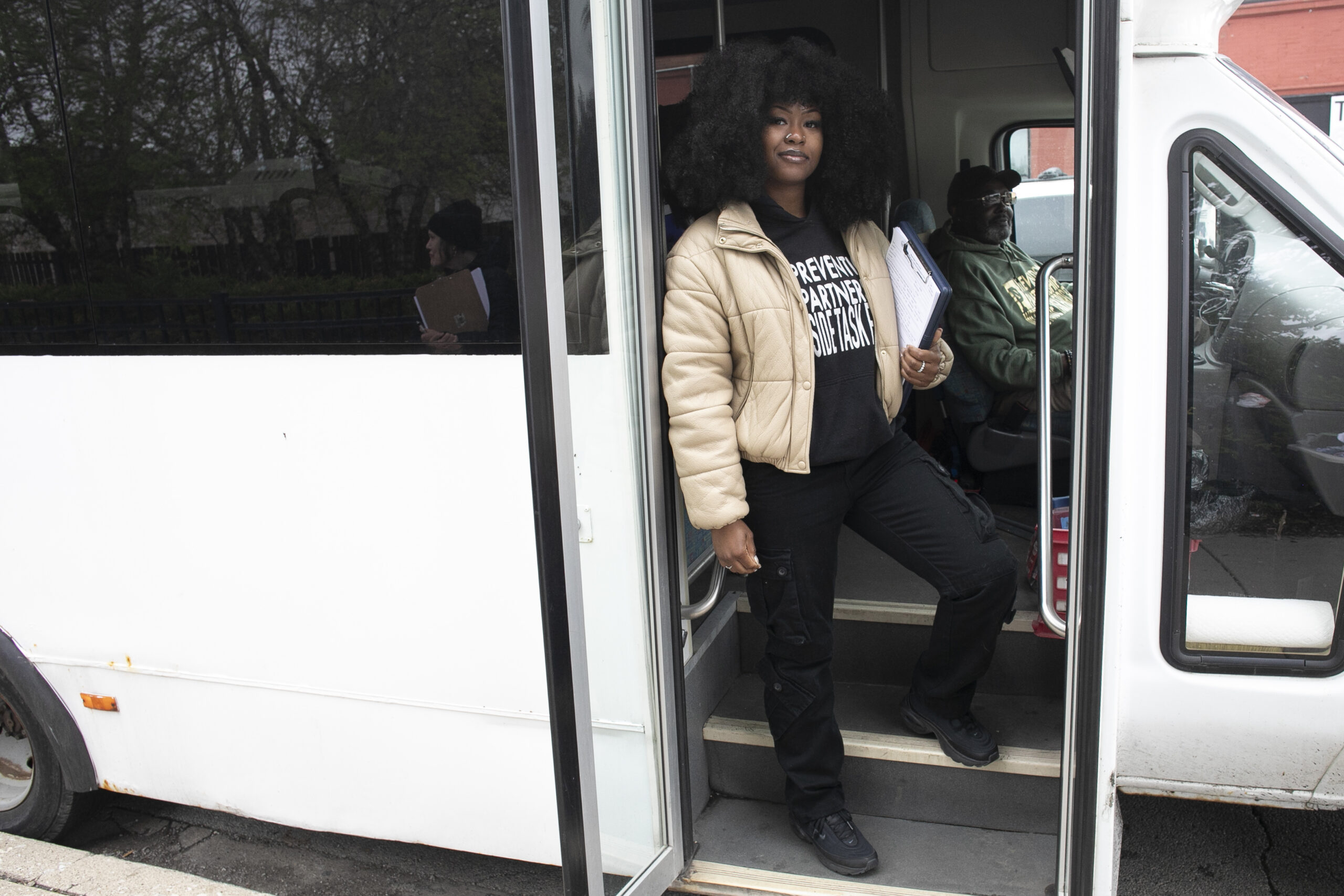 A woman with curly hair stands at the open door of a white bus, holding a clipboard and wearing a beige jacket and black hoodie. A man sits inside the bus.